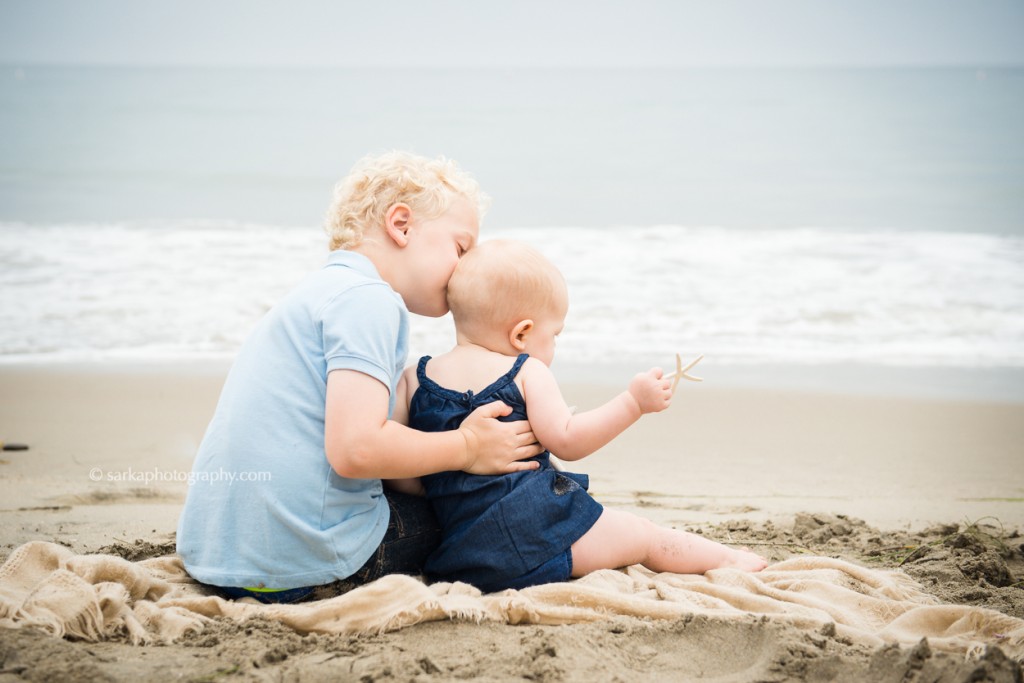 little boy kissing his younger sister sitting on a beach during their family photo session in Montecito Santa Barbara by sarkaphotography