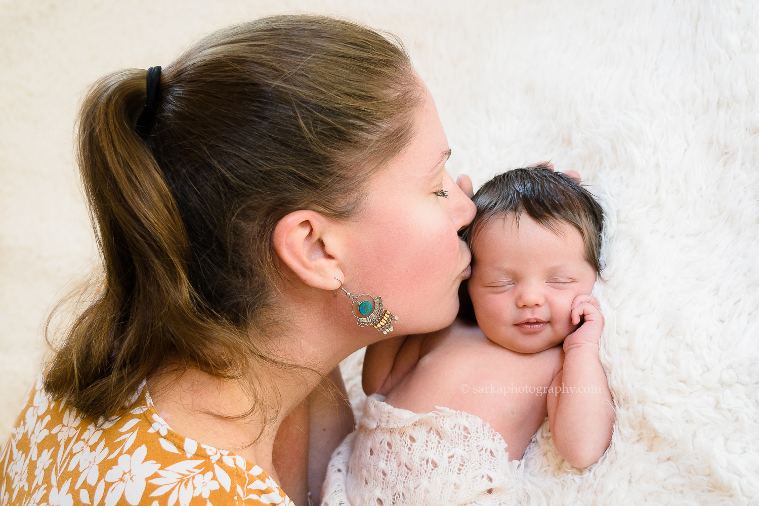 newborn baby girl softly smiling while her mom is kissing her cheek for their baby portrait